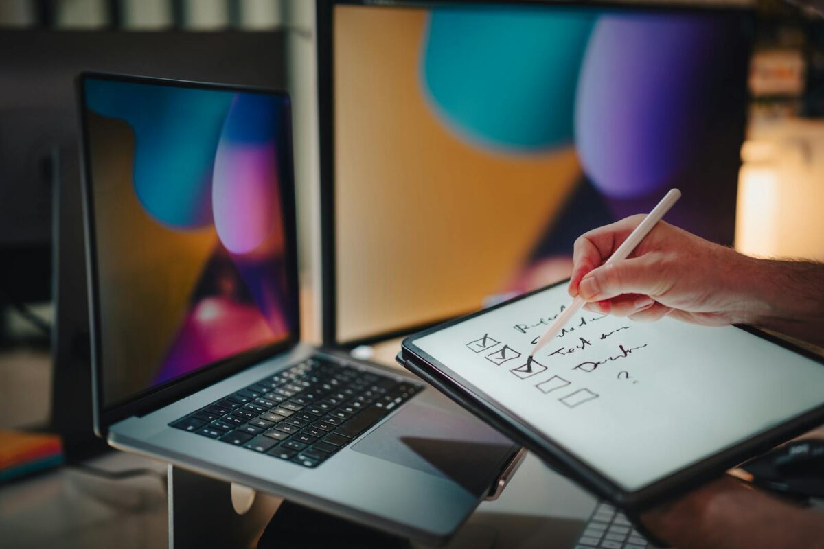 A hand writing on a tablet with stylus in a contemporary office setup featuring laptops and digital screens.