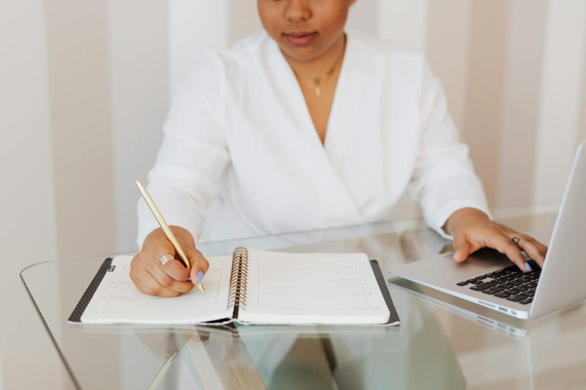 A focused professional woman planning her day with a notebook and laptop at a modern glass desk.