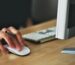 A hand using a wireless mouse at a modern desk setup with a computer and keyboard.