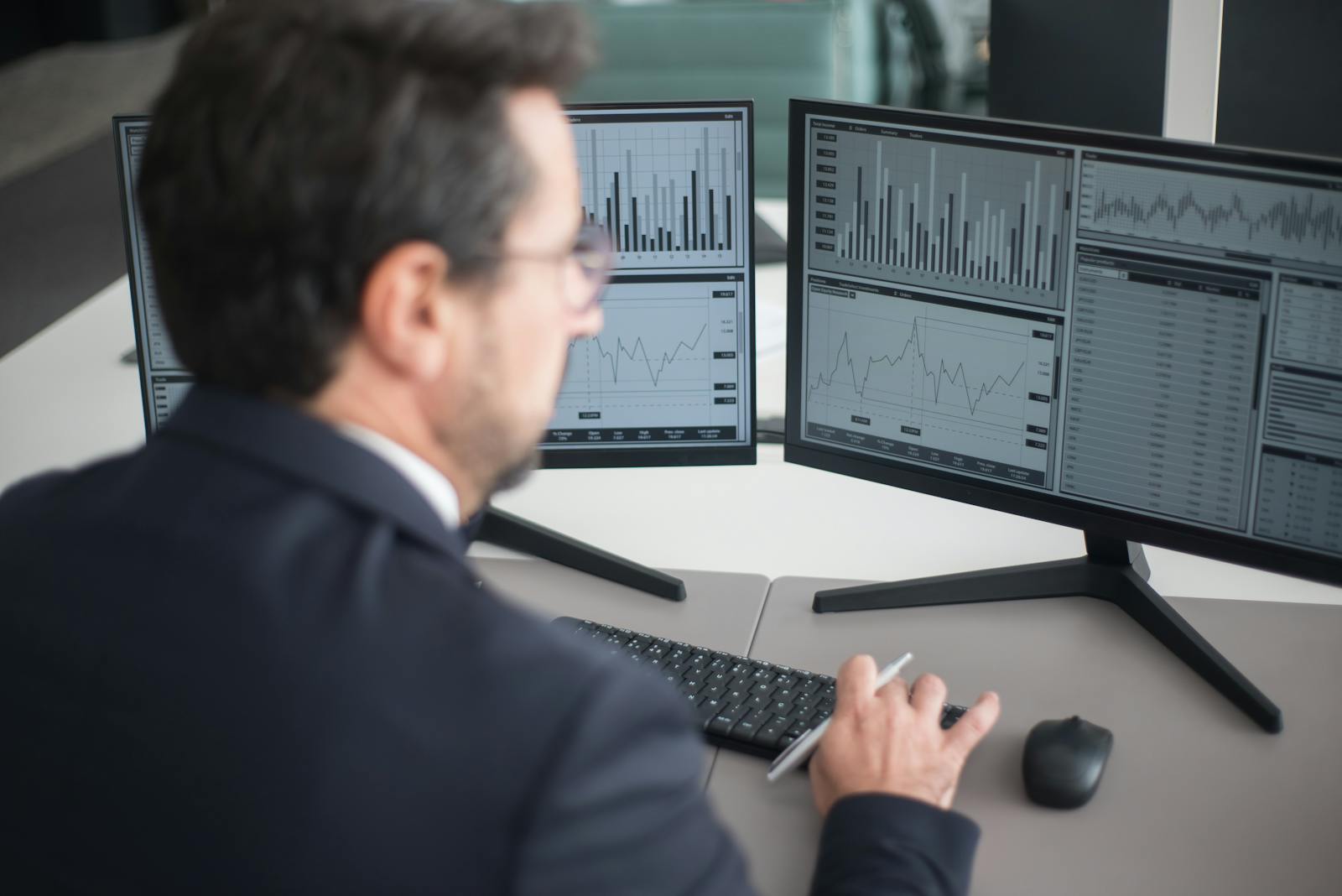 A businessman in a suit analyzing financial charts and graphs on two computer monitors indoors.