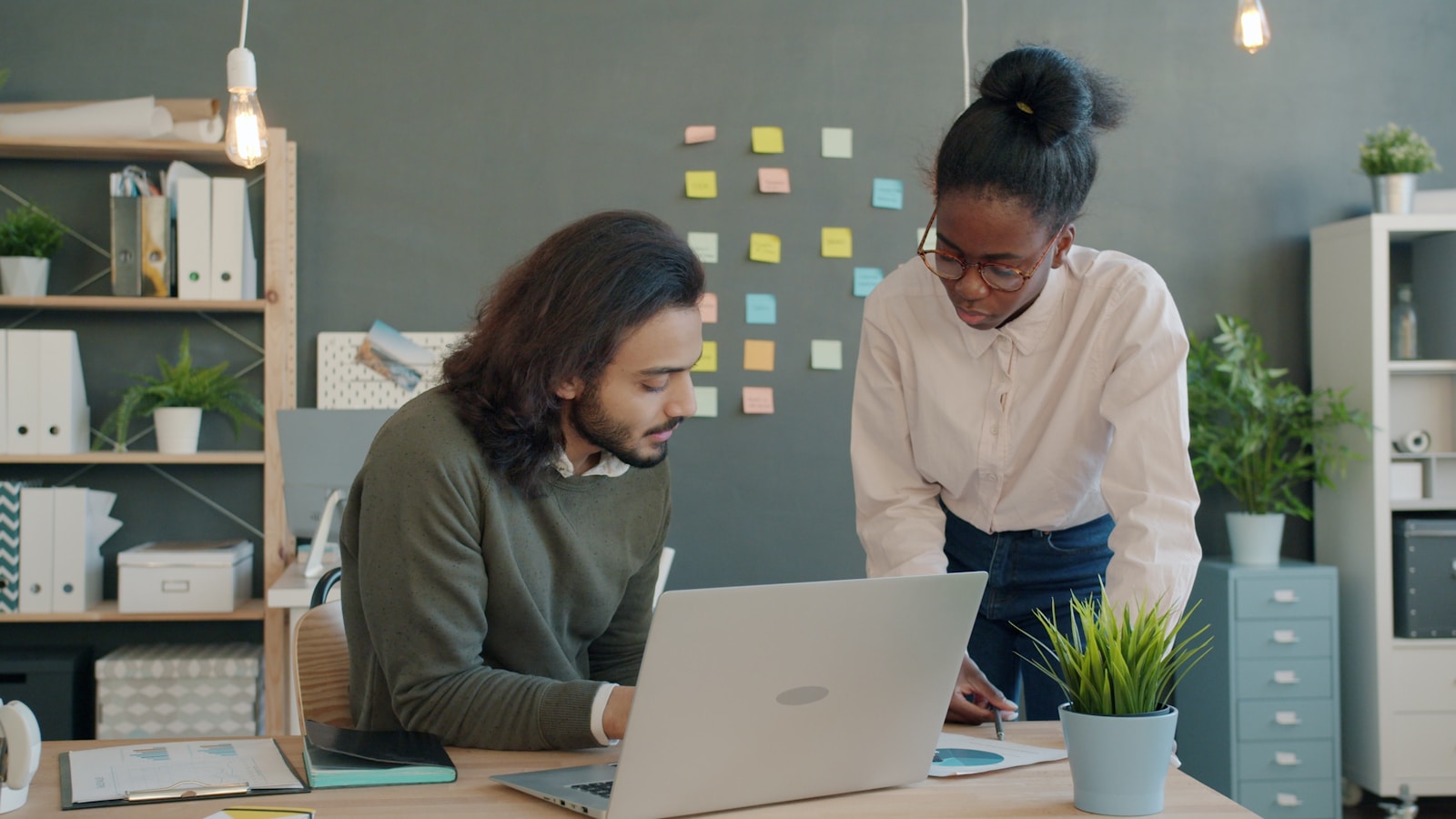 Sales team collaborating in a modern office, two colleagues reviewing data on a laptop while standing by a desk, with colorful sticky notes in the background for sales planning.