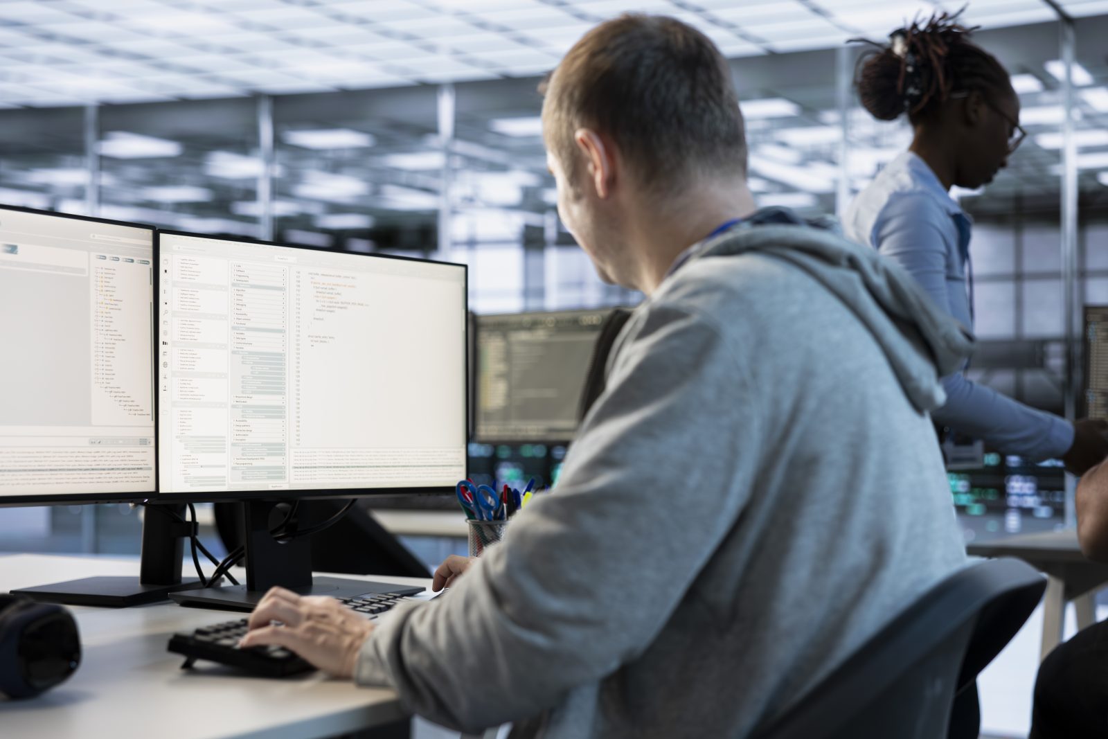 An engineer working at a dual monitor workstation with code on screen in a large office