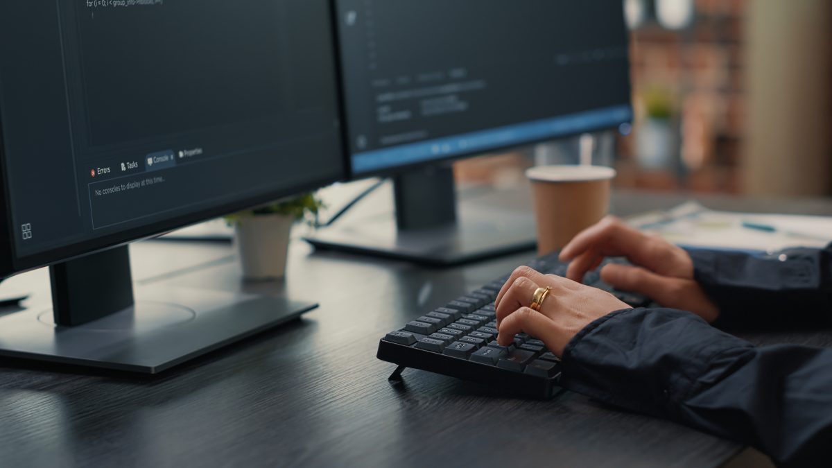 Close up of hands typing on a keyboard in front of two monitors displaying lines of code