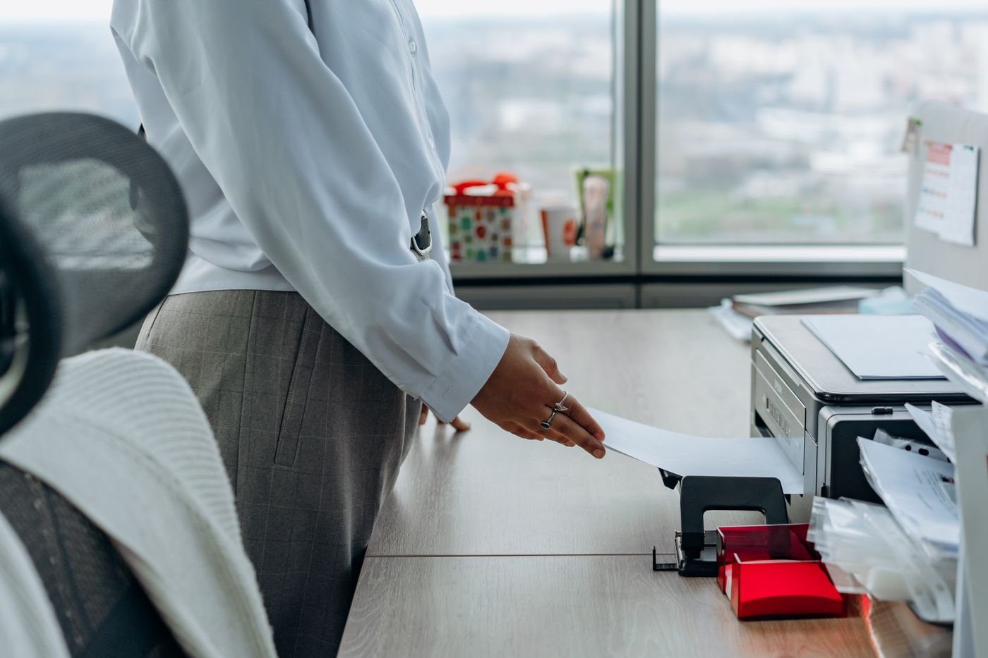 Office worker taking a freshly printed page from a desktop printer next to a window