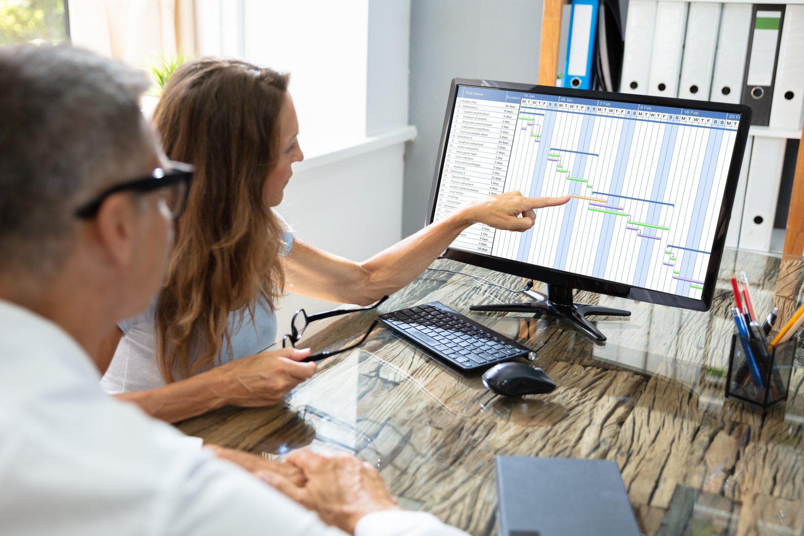 Two colleagues at a desk pointing at a Gantt chart of project tasks displayed on a monitor