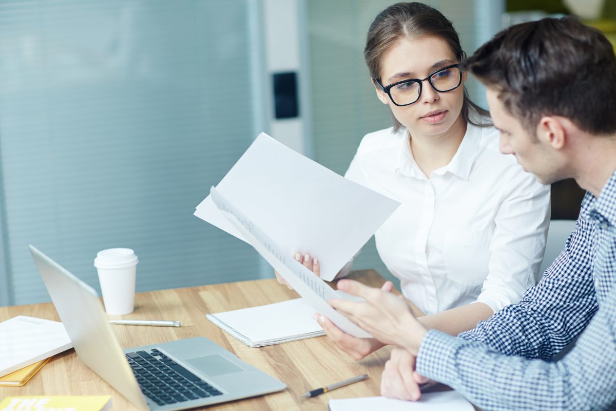 Two people going through a multi page printed contract at a meeting table with a laptop