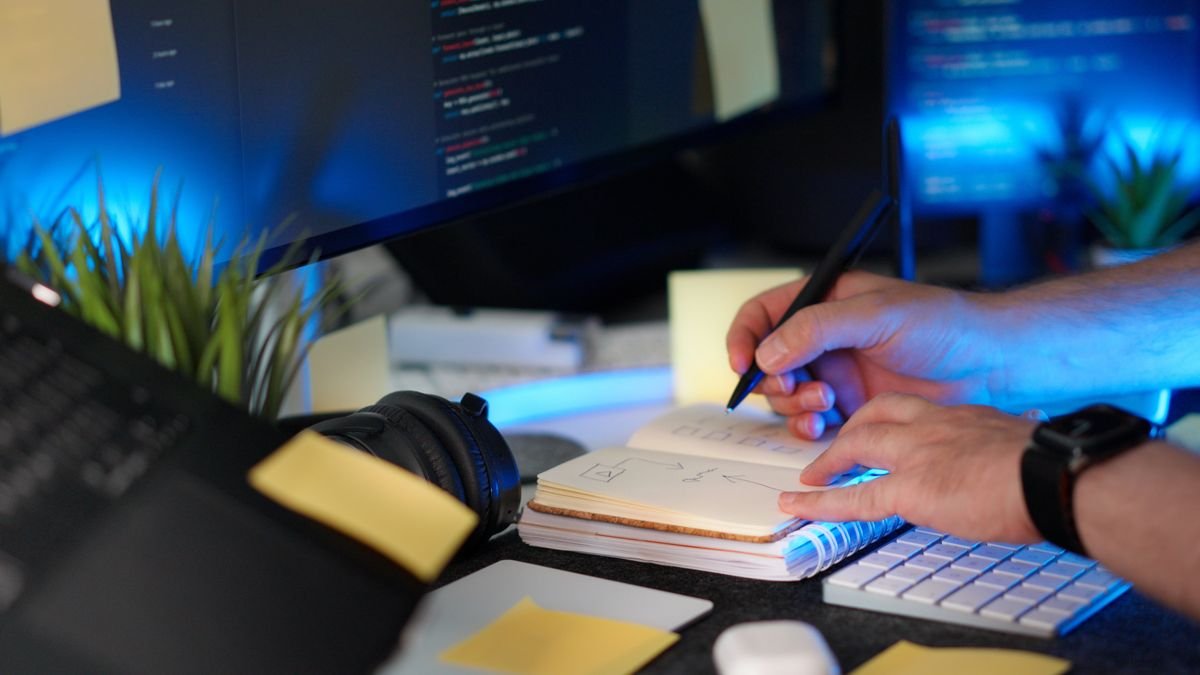 A person taking handwritten notes next to a laptop showing code, planning a workflow automation task