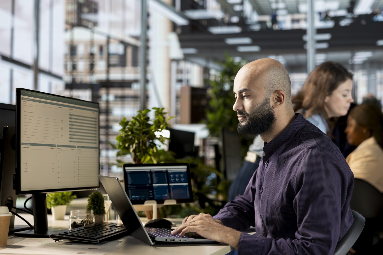 Man working at a two screen desk reviewing reports in an open plan office