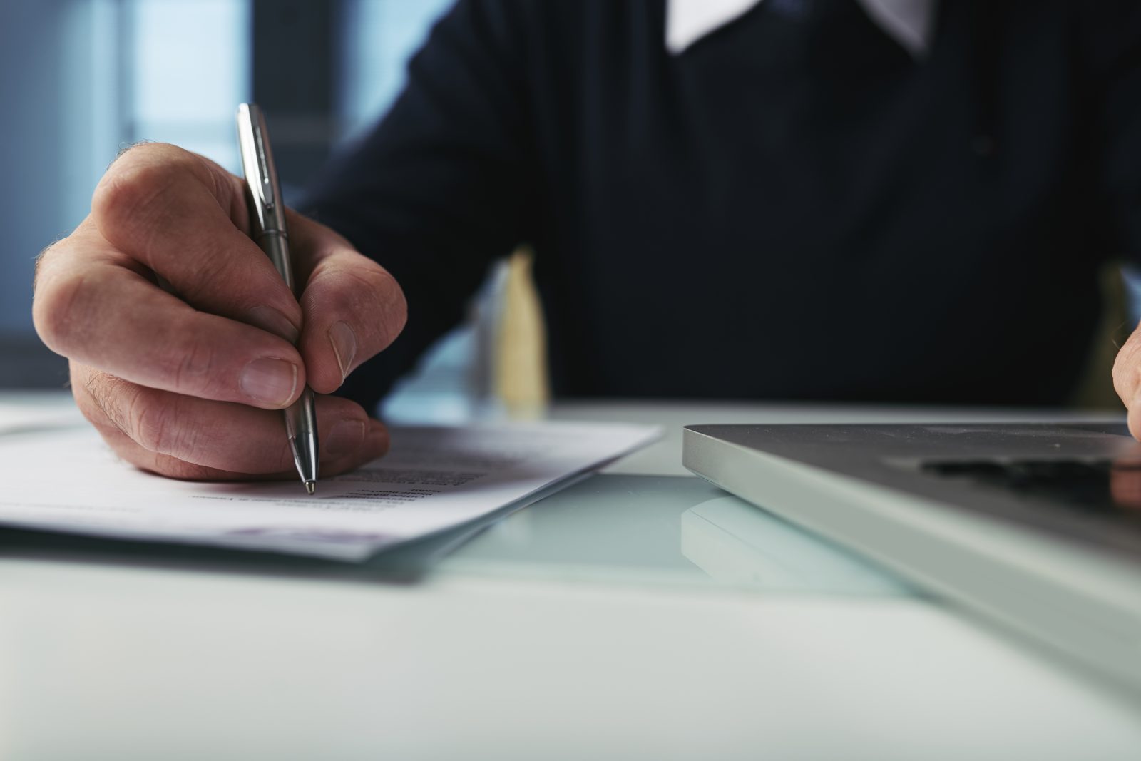 Close up of a person holding a pen over a printed contract page beside a laptop