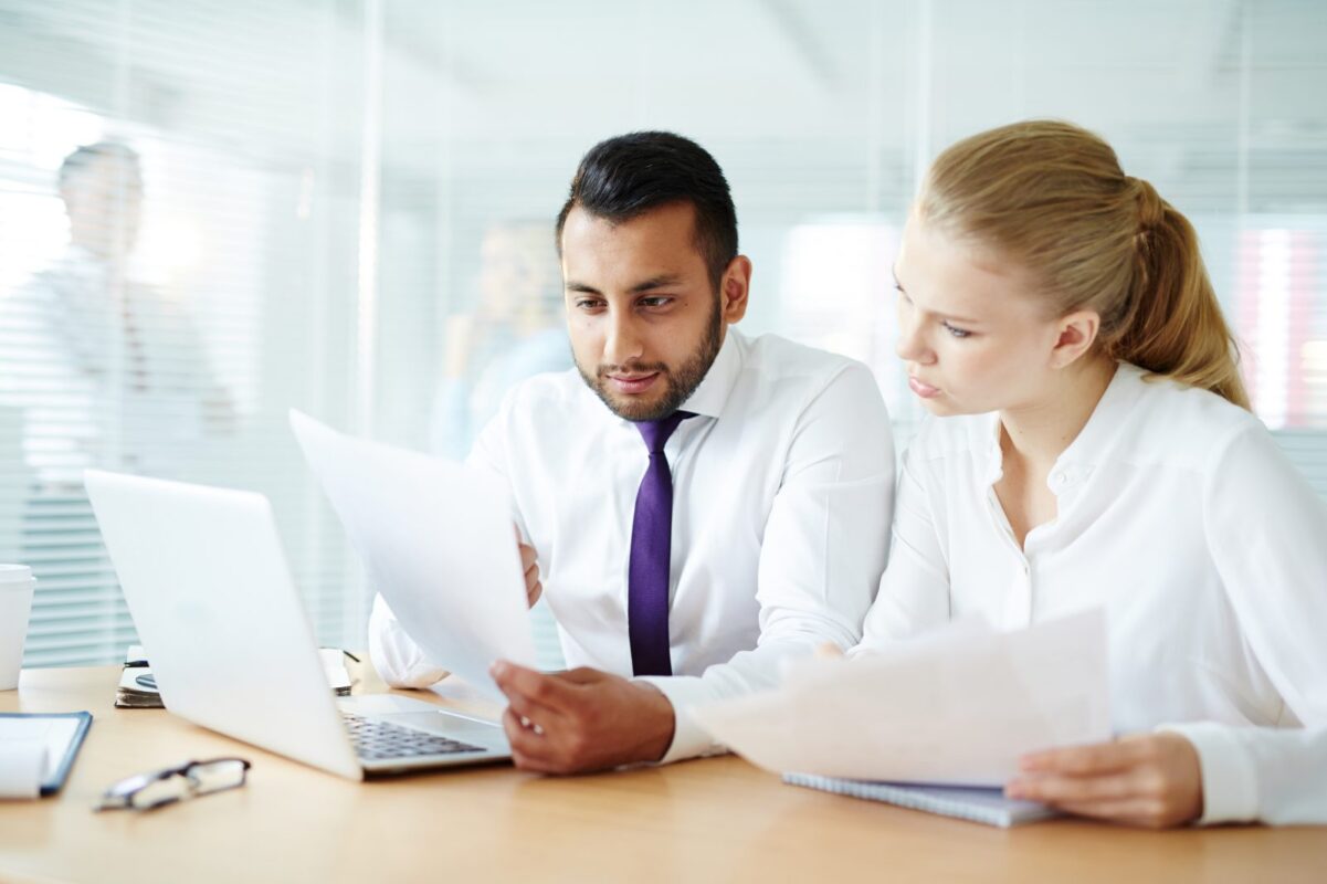Two business colleagues at a desk reading printed proposal pages next to an open laptop