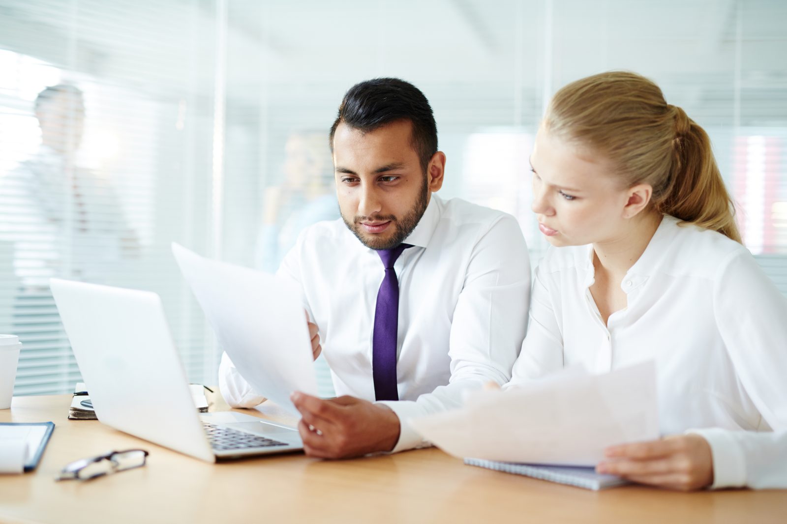 Two business colleagues at a desk reading printed proposal pages next to an open laptop